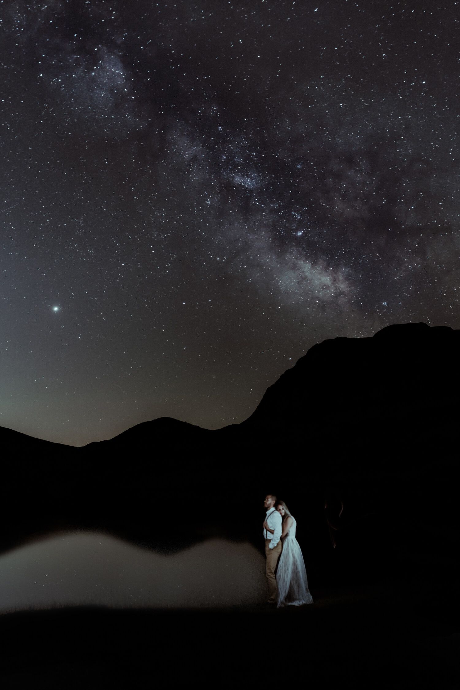 Le couple se tient au bord d'un lac de montagne dans les Pyrénées sous la voie lactée. Une astrophotographie réalisée par Gaétane Glize pendant leur séance photo de fiançailles.