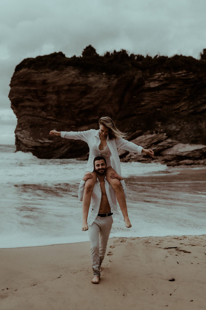 Un couple s'amuse sur la plage de Biarritz pendant leur séance photo fiançailles au Pays Basque. On y voit l'océan et les falaises.