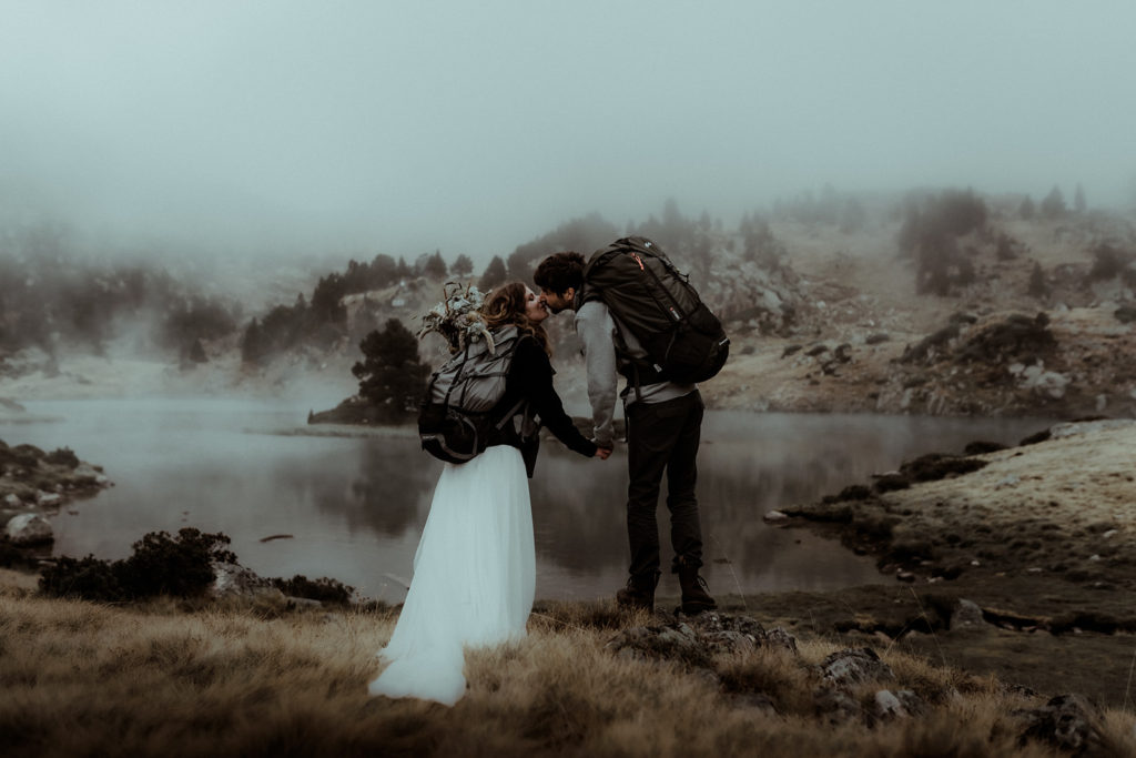 Photo d'un couple qui s'embrasse au bord d'un lac alpin dans les montagnes des Pyrénées. On y voit des sapins et des nuages bas. Ils sont en tenues de mariés et ont leur sac à dos de bivouac sur le dos. Photo prise lors de leur séance photo couple aventurière à l'occasion de leur elopement.