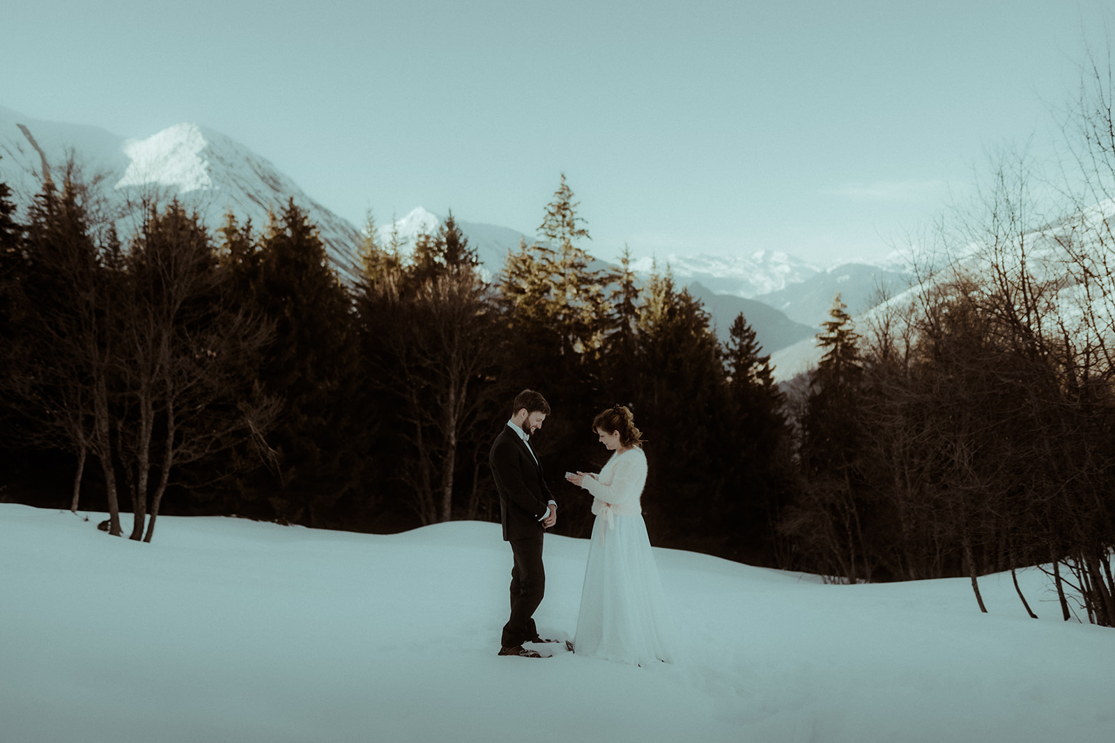 Un couple de mariés en costume et robe de mariée se tient au milieu des sapins et des montagnes enneigées. Ils ont fait une randonnée en raquette pour leur elopement dans les Pyrénées avec leur photographe Gaétane Glize.