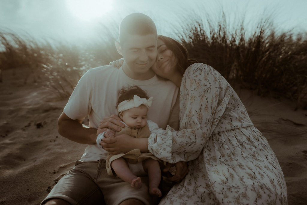 Une famille avec un nouveau-né est assise sur la dune au coucher du soleil à Seignosse dans les Landes. Ils partagent un moment de tendresse pendant leur séance photo bébé avec Gaétane Glize.