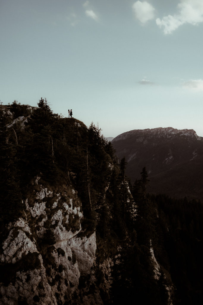 Un couple se tient sur une falaise dans les montagnes pendant leur séance photo de couple.