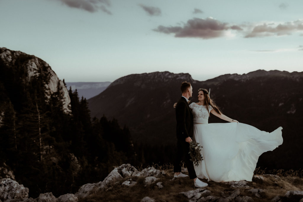 Un couple de mariés se tient au sommet d'une montagne dans les Pyrénées. La robe de la mariée vole au vent. Une photo capturée par Gaétane Glize, photographe elopement Pyrénées.