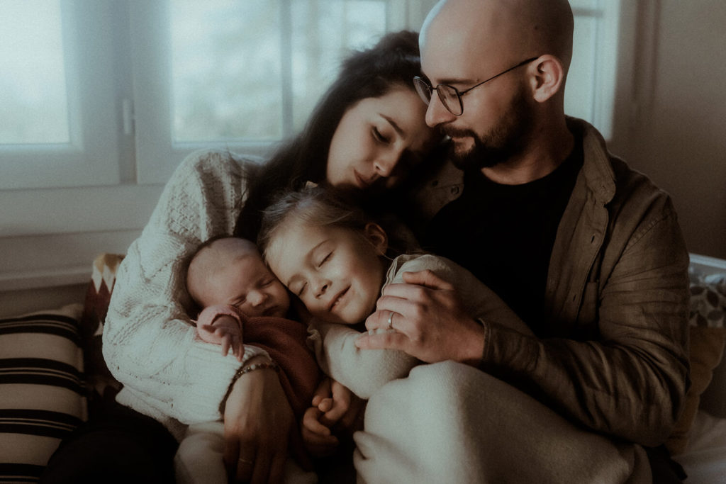Une famille composée d'une mère, d'un père, d'une fille et d'un nouveau-né se font un câlin sur le canapé. Un moment tendre saisi pendant leur séance photo nouveau-né en famille à Hossegor dans les Landes.