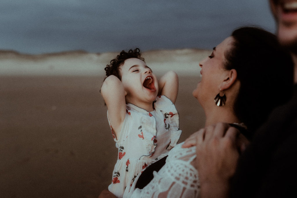 Une petite fille explose de joie, ce qui fait rire ses parents. La famille réalise sa séance photo sur la plage de Hossegor dans les Landes.