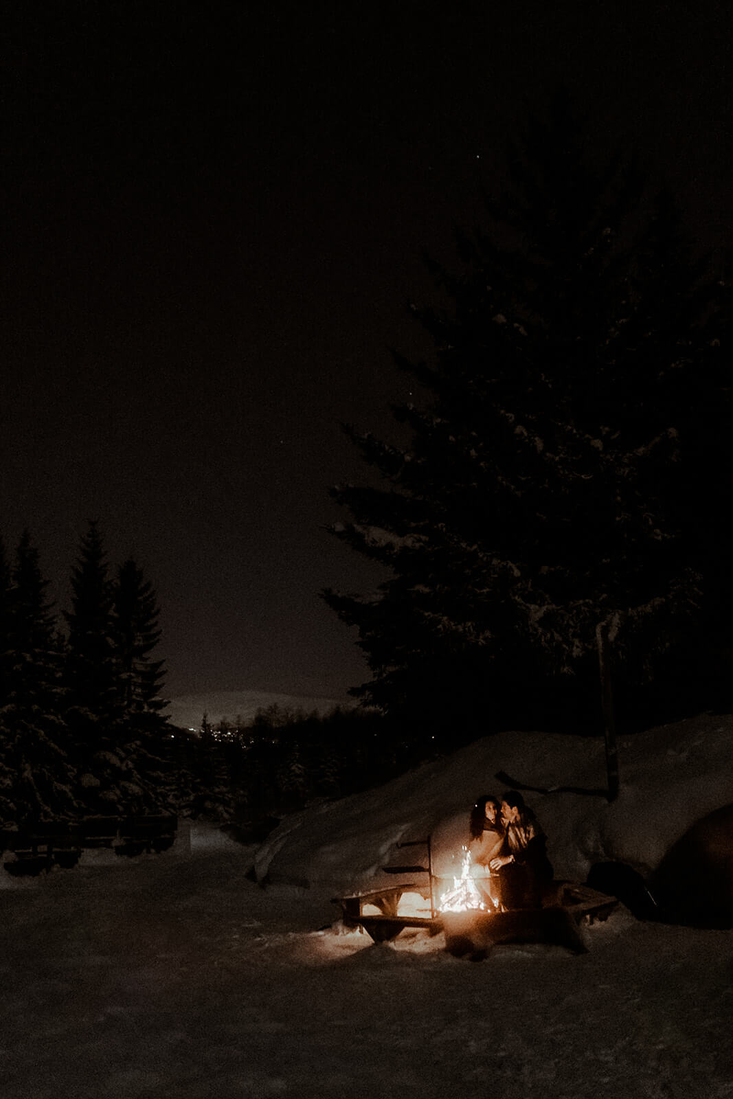 Un couple est assis au bord d'un feu de camp de nuit en montagne, ils sont emmitouflés dans un plaid et se font un câlin pendant leur séance photo de couple pour leurs fiançailles à Tromso en Norvège.