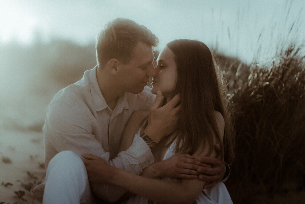 Un couple est assis dans les dunes de Seignosse, on voit la lumière dorée du coucher de soleil. Ils sont sur le point de s'embrasser pendant leur séance photo couple de fiançailles dans les Landes.