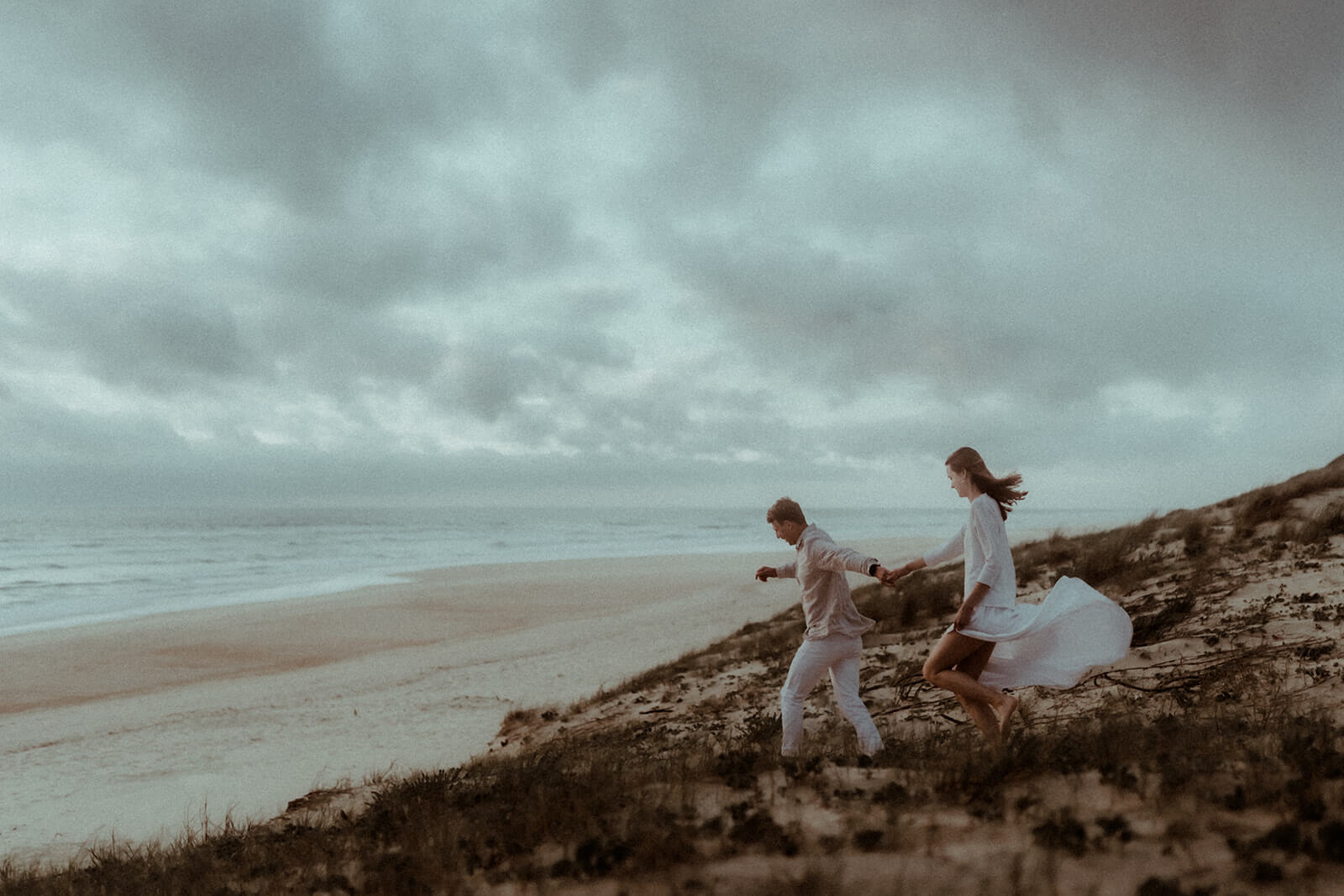 Un couple descend la dune de la plage de Seignosse en courant. Ils se dirigent vers l'océan. Un moment fun pendant leur séance photo de fiançailles dans les Landes.
