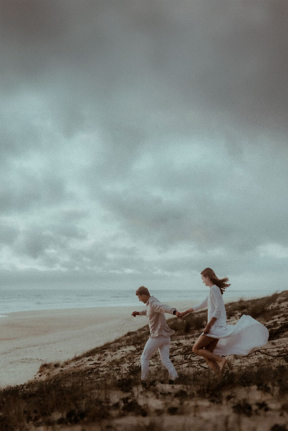 Un couple court main dans la main sur la dune d'une plage de Seignosse. On voit l'océan en fond et un ciel chargé de beaux nuages. Un moment fun pendant leur séance photo de fiançailles avec Gaétane Glize, photographe couple landes.