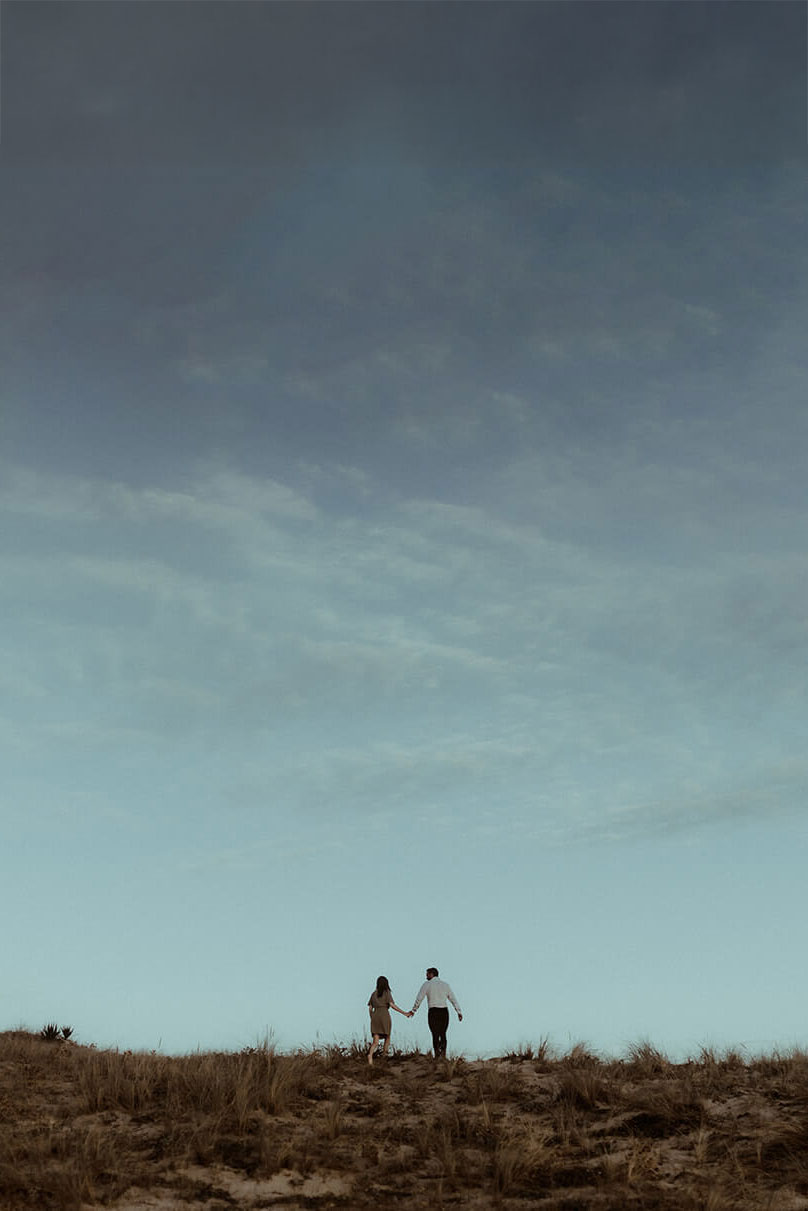 Un couple marche main dans la main sur la dune de la plage de Seignosse, le ciel est bleu. Leur séance photo de couple était à l'occasion de leurs fiançailles.