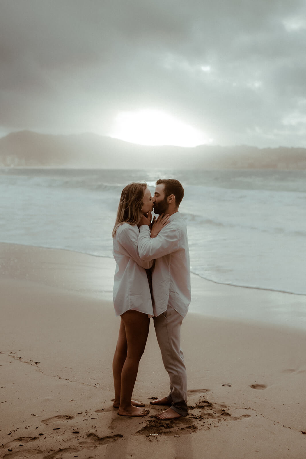 Un couple s'embrasse au coucher du soleil sur la plage de Biarritz, on voit les montagnes et l'océan en fond. Un moment saisi par Gaétane Glize, photographe couple landes et pays basque.