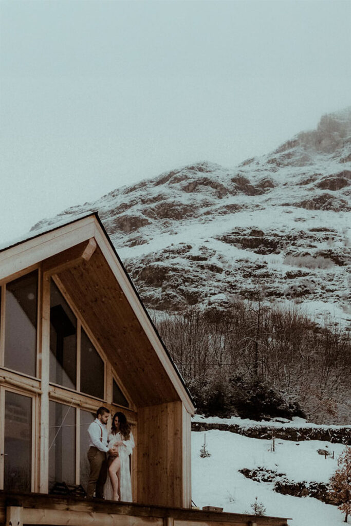 Un couple se tient devant une cabane en bois en montagne, la neige les entoure. Ils ont loué ce chalet à Gavarnie dans les Pyrénées pour leur séance photo de grossesse avec leur photographe Gaétane Glize.