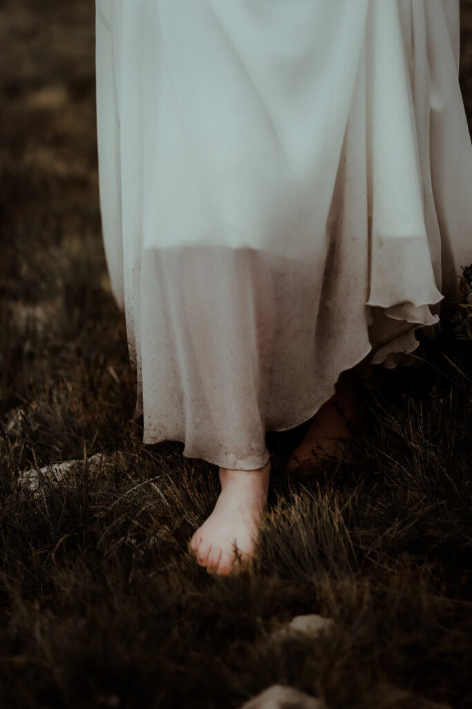 La mariée marche pieds nus dans l'herbe. Elle soulève sa robe de mariée au tissu fluide et transparent. Une mariée un brin sauvage le jour de son elopement dans les montagnes des Pyrénées.