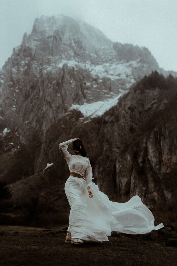 La mariée se tient au pieds d'une montagne enneigée. Sa robe vole avec le vent. Une photo artistique capturé par Gaétane Glize, photographe elopement Pyrénées.