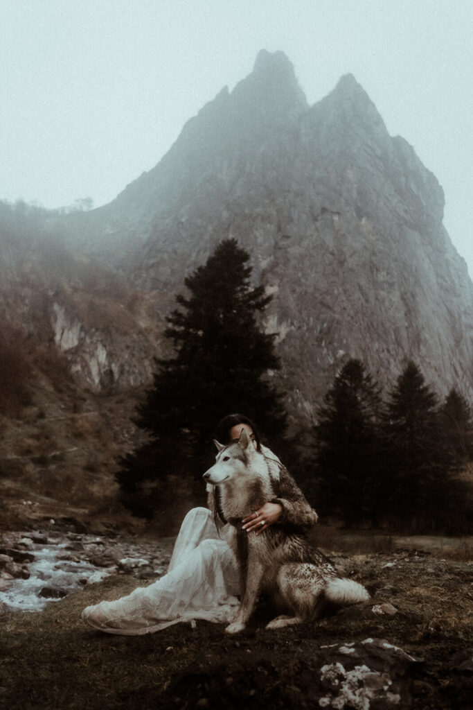 La mariée est assise avec son chien-loup croisé husky. Elle est au bord d'une rivière, au pied d'un pic montagneux. Un moment photographié pendant son elopement dans les Pyrénées.