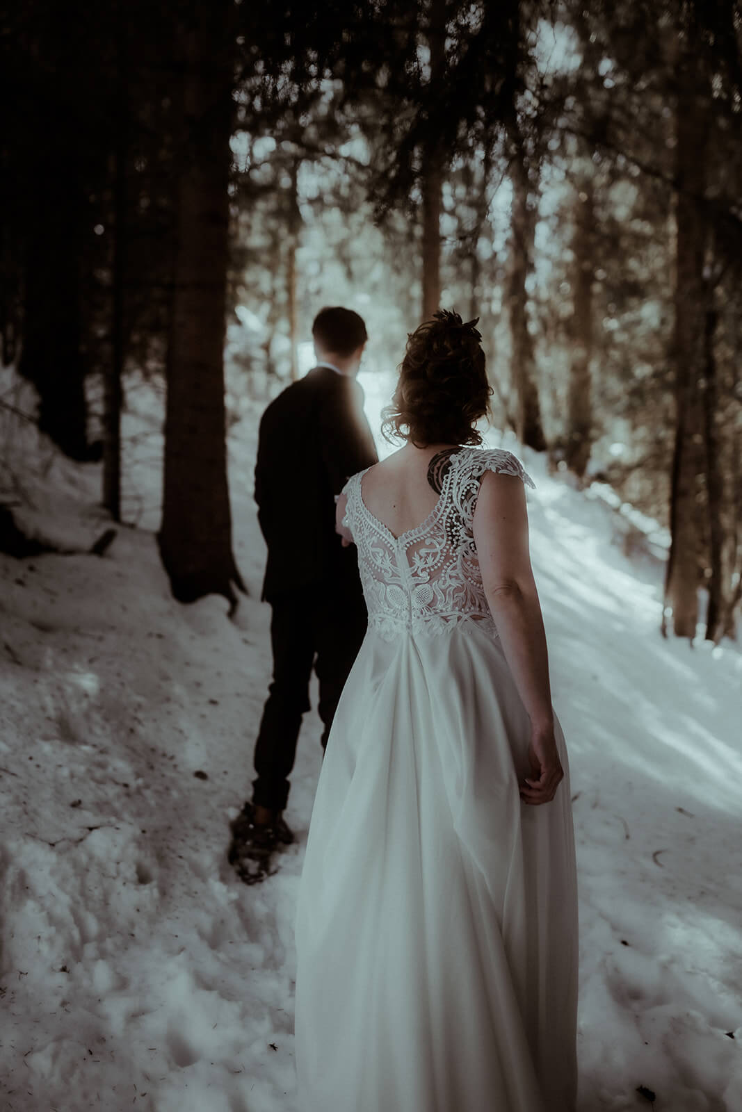 Le marié en costume marche devant et tient la main de sa femme en robe de mariée. Ils ont des raquettes aux pieds, sont entourés de sapins et de neige. Un elopement d'hiver en montagne dans les Pyrénées.